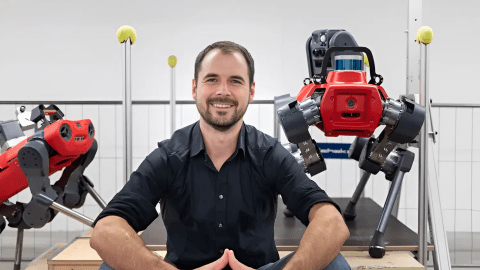 Marco Hutter sitting in a robotics lab with two advanced quadruped robots behind him. The image shows a modern robotics environment focused on autonomous systems, robot locomotion, and engineering innovation.