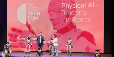 Two speakers stand on stage surrounded by humanoid and quadruped robots at the Davos Tech Summit. A large pink screen behind them displays the event title “Physical AI – Touching Intelligence” and the details “July 1–4, 2026, Davos Congress, www.davostechnosummit.com.”  The backdrop features a futuristic robot face and hand in shades of red and pink.